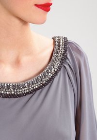 Šedá blůza s kulatým výstřihem ozdobená stříbrnými a černými korálky. Texturovaná látka vytváří jemné splývání kolem ramena.