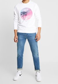 Sweat-shirt blanc avec un graphisme circulaire représentant des palmiers et des couleurs de coucher de soleil, assorti à un jean bleu et des baskets blanches.