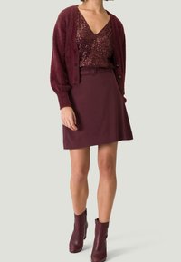 Haut en sequins bordeaux superposé avec un cardigan en fausse fourrure bordeaux, associé à une jupe bordeaux et des bottines assorties. Tissus lisses et texturés.