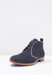 Bottines en daim bleu foncé avec un bout arrondi, des coutures grises et un talon en bois présentant un accent bleu à la base. Design à lacets.
