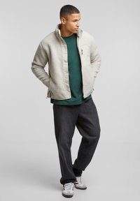 Gaišs pelēks fliša jakas mētelis ar augstu apkakli, ar rāvējslēdzējam kabatām, valkāts virs tumši zaļas krekla un apvienots ar tumšiem biksēm un pelēkiem sporta apaviem.