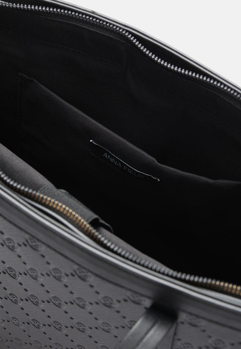 Interior de una bolsa tote negra texturizada con un forro de tela suave, que cuenta con un cierre de cremallera y una etiqueta que dice "ANNA FIELD."