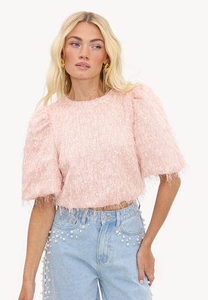 Femme blonde portant un haut texturé rose à manches courtes avec des sequins et un jean taille haute bleu clair orné de perles.