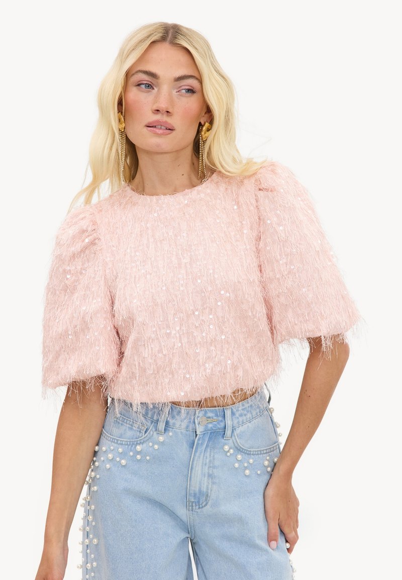 Femme blonde portant un haut texturé rose à manches courtes avec des sequins et un jean taille haute bleu clair orné de perles.
