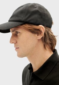 Gorra de béisbol de algodón negro con visera estructurada, que presenta agujeros de ventilación laterales y un diseño ajustado. Se lleva con una camiseta polo oscura.