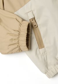 Leichte, beige Jacke mit einer weichen Textur. Verfügt über elastische Bündchen, eine mit Reißverschluss verschließbare Tasche und ein kontrastierendes beiges Oberteil.