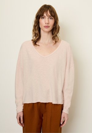 Haut rose clair à manches longues avec un décolleté en V, des bords côtelés et un ourlet brut. Porté avec un pantalon brun ample et texturé.
