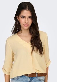 Lichtgele blouse met een V-hals en driekwart mouwen, uitgevoerd in een gestructureerde stof en een losse pasvorm. Gecombineerd met een bruine riem en blauwe denim.