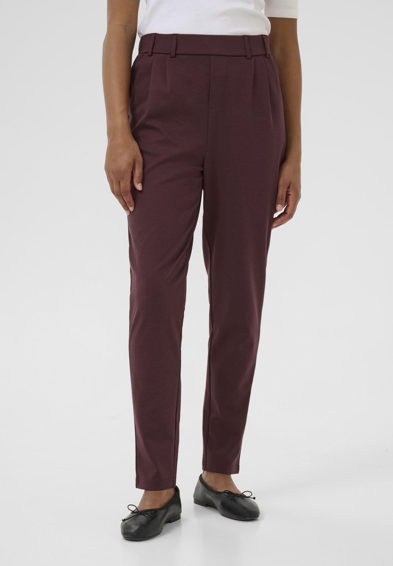 Pantaloni marrone scuro a vita alta con taglio dritto, con pieghe frontali e passanti per cintura. Tessuto liscio e lunghezza alla caviglia. Scarpe nere.
