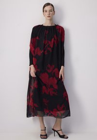 Abito lungo nero a fiori con motivi floreali rossi; presenta un colletto arricciato e un soprabito in tessuto trasparente. Indossato con tacchi a strappo neri.