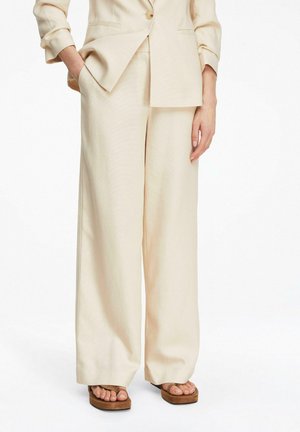 Frau trägt cremefarbene Hosen mit weitem Bein und passend dazu Blazer, eine Hand in der Tasche, kombiniert mit braunen Plateausandalen.