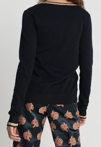 Marineblaue Strickjacke mit Rundhalsausschnitt und gerippten Bündchen, versehen mit einem goldenen Streifen am Kragen, kombiniert mit gemusterten Hosen.