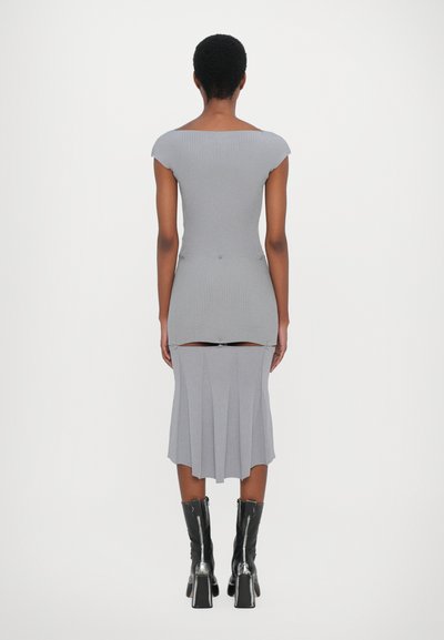 Vestido gris de canalé con mangas cortas, cuerpo ajustado y falda con pliegues. Presenta cierres a presión para un ajuste ajustable. La modelo lleva botas negras hasta el tobillo.