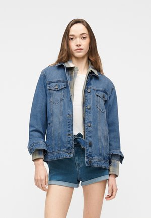 Jeune femme portant une veste en jean superposée, une chemise à carreaux, un haut blanc et un short en jean retroussé, debout devant un fond uni.