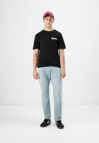 Zwarte katoenen t-shirt met kleine print, lichtblauwe jeans en zwarte sneakers. Rode pet. Staand op een effen witte achtergrond.