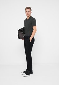 Polo grigio a maniche corte, pantaloni neri slim-fit e sneaker nere con suola bianca. Il modello tiene una giacca in un braccio.