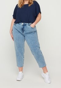 Jeans mom blu in denim con vita alta e gamba tapered, dotati di chiusura con bottone e tasche frontali. Abbinati a una maglietta larga di colore blu navy.