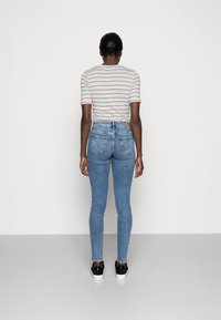 Jeans ajustados de mezclilla azul con cintura alta, dos bolsillos traseros y un tono claro, combinados con una camiseta de rayas de manga corta y zapatillas negras.