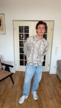 Fiatal férfi világos mintás pulóverben, kék farmernadrágban és fehér tornacipőben, kezeit a hát mögött tartva, egy fából készült padlójú szobában áll.