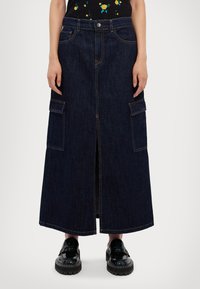 Lange, donkere denimrok met zijcargo pockets, een split aan de voorkant en contrasterende stiksels, gecombineerd met zwarte plateauschoenen.