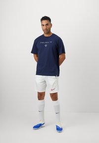 Mand står med hænderne bag ryggen iført navy Chelsea FC t-shirt, hvide Nike shorts, hvide sokker og blå fodboldsko mod en hvid baggrund.