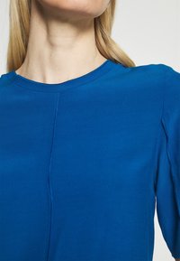 Niebieska bluzka z długim rękawem i okrągłym dekoltem, charakteryzująca się gładką teksturą oraz widocznymi szwami na przodzie i rękawach.