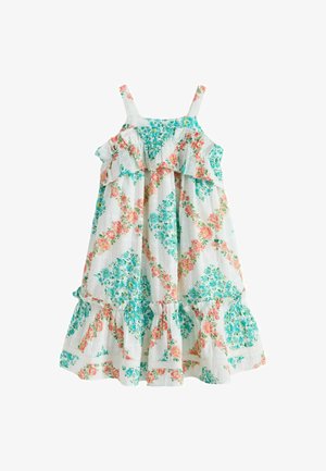 Mladé dievča sa usmieva, nosí biele bezrukávové kvetované šaty s volánmi, stojí pred jednofarebným béžovým pozadím.