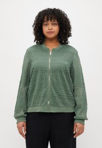 Veste bomber en dentelle vert clair avec fermeture éclair. Présente des motifs complexes, des poignets et un ourlet côtelés. Tissu léger et respirant.