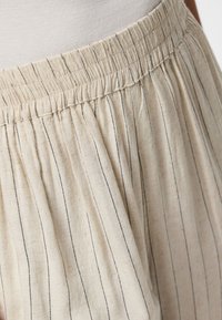 Jupe ou pantalon beige avec de fines rayures verticales noires et une taille élastique, porté avec un top blanc.