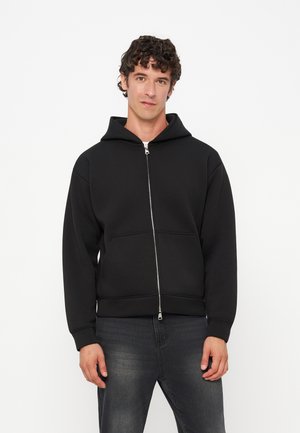 Jeune homme aux cheveux bouclés portant un sweat à capuche noir zippé et un jean foncé, debout devant un fond blanc uni.