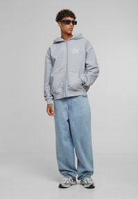 Siva zip-up dukserica sa prednjim džepovima, sa bijelim tekstom. Uparen sa širokim svijetloplavim farmerkama i sivim patikama s naglascima.