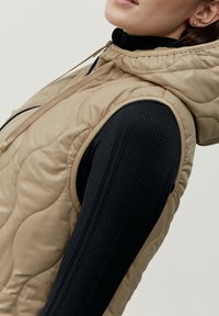 Beige kviltad ärmlös väst med huva, med en slät yta och ett svart långärmat baslager med ribbat mönster.