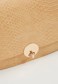 Béžová texturovaná kožená taška se zlatým kulatým zámkem na jednoduchém bílém pozadí.