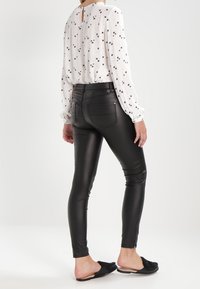 Pantaloni in ecopelle neri con una vestibilità slim, caratterizzati da una texture liscia e tasche posteriori. Abbinati a una blusa bianca a maniche lunghe, ampia, con motivi floreali.