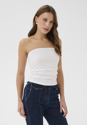 Femme aux longs cheveux bruns portant un haut blanc sans bretelles et un jean taille haute bleu foncé, debout devant un fond clair uni.