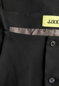 Närbild av svart plagg med brunt foder, två svarta knappar och en gul etikett med texten "JJXX" på insidan av midjebandet.