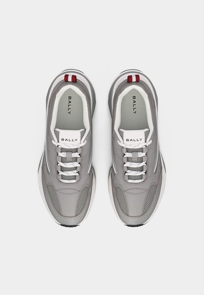 Ζευγάρι γκρι και λευκά αθλητικά παπούτσια Bally με λευκά κορδόνια, λεπτομέρειες από δίχτυ και κόκκινες ραβδωτές ετικέτες στη φτέρνα, εμφανισμένα από πάνω σε λευκό φόντο.