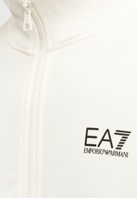 Λευκό τζάκετ με φερμουάρ από λείο ύφασμα, με ψηλό γιακά και μαύρο λογότυπο "EA7 Emporio Armani" στο στήθος.