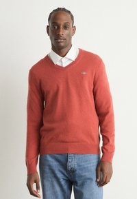 Pull rouge en V réalisé en tissu tricot doux, avec des poignets et un ourlet côtelés, porté sur une chemise blanche à col avec un jean bleu.