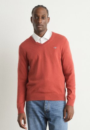 Roter V-Ausschnitt-Pullover aus weichem Strickmaterial, mit gerippten Bündchen und Saum, getragen über einem weißen Hemd mit Kragen und blauen Jeans.