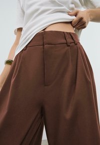 Pantaloni a pieghe marroni realizzati in un tessuto morbido, con vita alta e tasche laterali, abbinati a una maglietta bianca.
