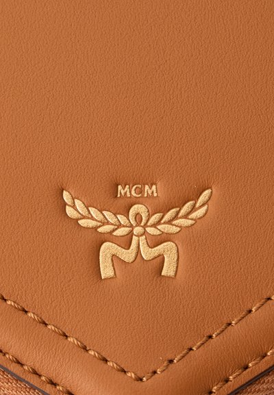 Surface en cuir fauve avec logo MCM doré en relief présentant des ailes stylisées et des feuilles de laurier au-dessus de coutures détaillées.