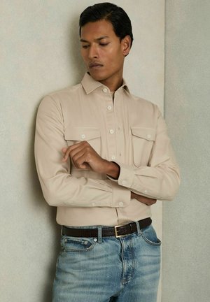 Chemise beige à manches longues avec deux poches poitrine, design à boutons et texture lisse, associée à un jean bleu et une ceinture noire.