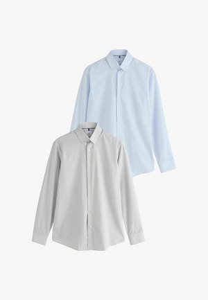 Giovane in piedi che indossa una camicia grigio chiaro con bottoni, pantaloni neri, cintura marrone e scarpe di pelle marrone su sfondo bianco uniforme.