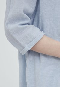 Ljusblå, texturerad tygärm med rullad manschettdetalj. Mjuk, andningsbar material med subtilt vävd mönster och mjuk drapering.