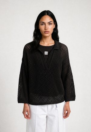 Frau mit langen dunklen Haaren trägt einen schwarzen grob gestrickten Pullover über einem schwarzen Top und weißen Hosen, steht vor einem schlichten hellen Hintergrund.