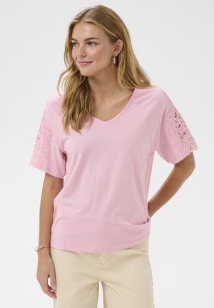 Moteris dėvi šviesiai rožinę V formos kaklo palaidinę su nėrinių detalėmis trumpomis rankovėmis, smėlio spalvos kelnes, auksinius lankelio formos auskarus ir apyrankę, pozuojanti prieš baltą foną.