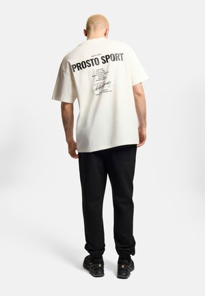 Mężczyzna z ogoloną głową nosi białą luźną koszulkę z napisem "PROSTO SPORT", czarne spodnie i czarne trampki, stoi tyłem.