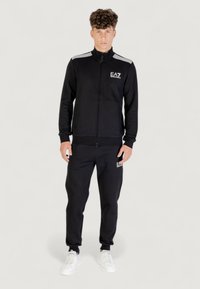 Chándal negro con chaqueta de cremallera y pantalones, que presenta un logotipo blanco y acentos a rayas, hecho de tela suave. Zapatillas blancas lisas.