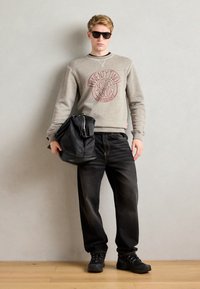 Grå hettegenser med rødt logo, svarte jeans, og svarte joggesko. Mann som holder en svart lærveske, bruker solbriller, står på tregulv.